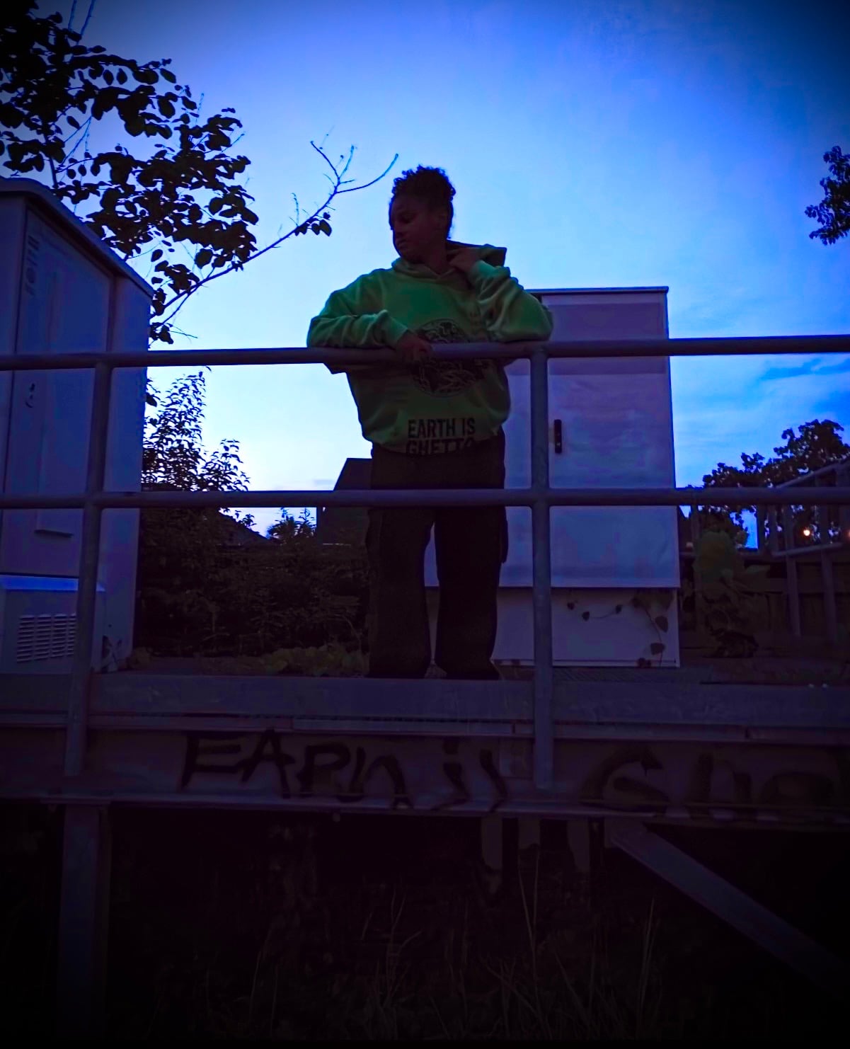 Campaign imagery: a person in a neon-green Earth Is Ghetto hoodie at a railing in blue dusk light.