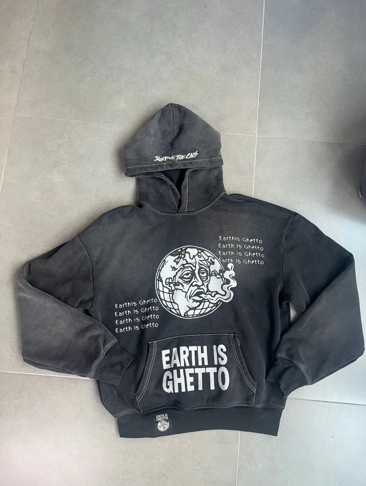 Sun-faded black hooded sweatshirt laid flat, front view: a globe graphic of a tired, smoking Earth with stacked "Earth Is Ghetto" text lines on either side, a kangaroo pouch pocket printed "EARTH IS GHETTO", and a woven label at the hem.